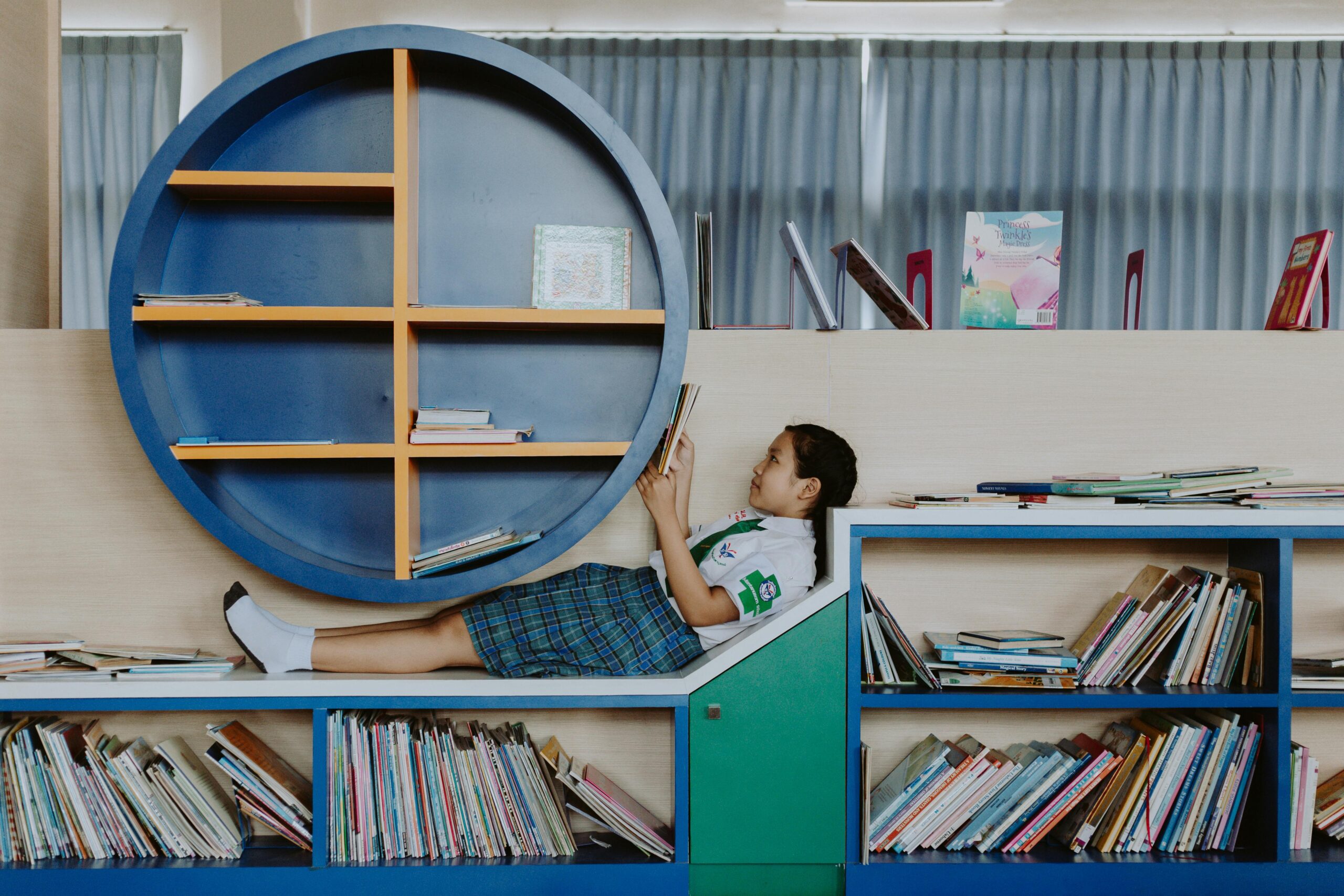 A young girl in a school uniform relaxes and reads in a modern library setting.